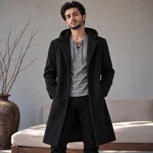 May include: A long, black, hooded overcoat with a structured collar and long sleeves, worn over a grey, short-sleeved shirt featuring a V-neckline with black lace-up detailing, paired with black trousers.