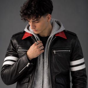 May include: A black leather jacket featuring a bright red collar, two white horizontal stripes on each sleeve, silver-toned metal zippers on the chest and cuffs, and two front pockets, worn over a light gray hooded sweatshirt with a drawstring.