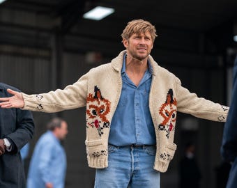 以下が含まれることがあります： クリーム色のチャンキーニットのジップアップカーディガンで、前身頃の両側に目立つキツネのモチーフがあしらわれています。ショールカラー、リブ編みの袖口と裾が特徴で、ライトブルーのボタンダウンシャツとライトブルーのデニムジーンズの上に着用されています。