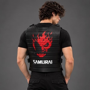 May include: A black tactical vest featuring horizontal ribbed panels and side adjustment straps. The back displays a large, vibrant red graphic of a stylized face with flames, positioned above the white text SAMURAI printed in a bold, block font.