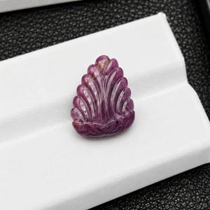 Peut inclure: Une pierre précieuse de couleur magenta profond sculptée en forme de goutte avec une série de crêtes verticales en éventail rayonnant de la base vers le sommet pointu. La pierre présente une surface polie et brillante avec des variations naturelles.