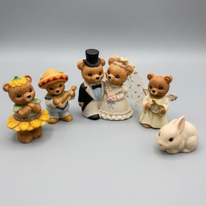 Puede incluir: Una colección de cinco figuras de cerámica que incluye cuatro osos de peluche marrones y un conejo blanco. Los osos incluyen uno vestido de girasol, uno con sombrero tocando la guitarra, una pareja de novios y un oso ángel con un arpa.