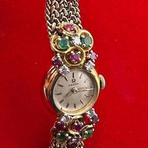 Puede incluir: Un reloj de pulsera Omega con esfera redonda plateada, manecillas e índices dorados. La caja y la pulsera tejida de oro están adornadas con grupos de rubíes, esmeraldas y diamantes redondos dispuestos en un patrón floral decorativo.