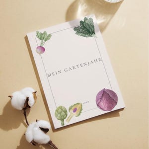 May include: A rectangular planner with a cream cover featuring the text MEIN GARTENJAHR and 2026. The cover is decorated with watercolour illustrations of a radish, leafy greens, two artichokes, and a purple cabbage, all within a thin black border.