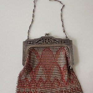 May include: A metal mesh handbag featuring a repeating pattern of small silver and coral-colored squares. The bag has an ornate, engraved silver-tone metal frame with a kiss-lock closure and a thin, delicate silver-tone chain shoulder strap.