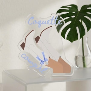 May include: A clear acrylic standee featuring an illustration of two white cowboy boots with brown heels and a light blue bow tied to one boot. The text Coquette is printed in light blue above the boots, and the word Cowgirl is printed below.