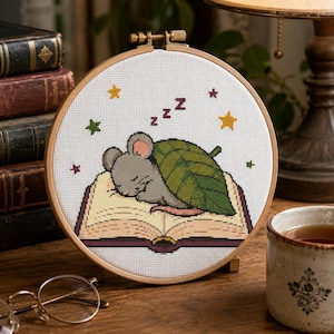 May include: A circular wooden embroidery hoop containing a cross-stitch pattern of a grey mouse sleeping on an open book while covered by a large green leaf. The design includes small colorful stars and the letter z repeated three times in dark purple.