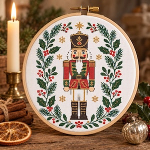 May include: A circular wooden embroidery hoop containing a cross-stitch pattern of a nutcracker figure centered within a wreath of green pine branches and red berries. The design includes small gold star accents on a white fabric background.