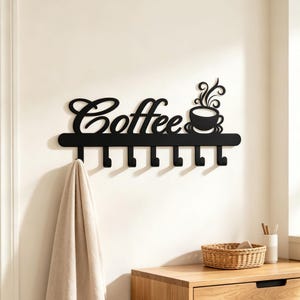 May include: A black wall-mounted metal rack featuring the word Coffee in cursive script, a graphic of a steaming cup, and seven downward-facing hooks. A beige towel hangs from the first hook on the left side of this functional wall organizer.