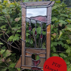 Op de afbeelding: Een hangend glas-in-lood ornament in de vorm van een guillotine, voorzien van amberkleurige glaspanelen, een reflecterend spiegelgedeelte en dieprode glazen accenten in het blad en de voet, opgehangen aan een zwarte metalen ketting.