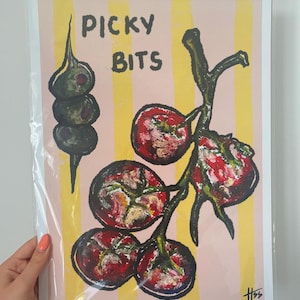 May include: A rectangular art print featuring a light pink background with vertical yellow stripes. The illustration depicts a skewer of three green olives and a cluster of red tomatoes on the vine. The text PICKY BITS is printed in black.