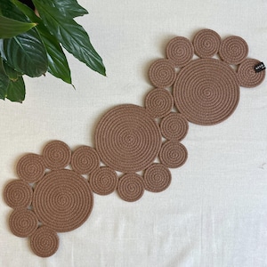 May include: A brown table runner constructed from multiple circular rope coils of varying sizes arranged in an organic, flowing pattern. The piece features a small black fabric tag on the upper right edge with the text KEYD printed in white.