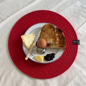 Pode incluir: Um lugar americano redondo de corda vermelha com uma etiqueta preta com a inscrição KEYD HANDMADE. Sobre o lugar americano, um prato branco com borda azul contém duas fatias de pão torrado, um ovo cozido num suporte de metal, queijo, manteiga e compota.