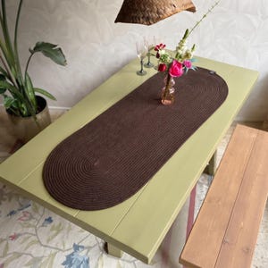 May include: An oval-shaped dark brown braided table runner made of thick cord, featuring a continuous spiral pattern. The runner is centred on a light green wooden table, providing a textured surface for a glass vase with colourful flowers.