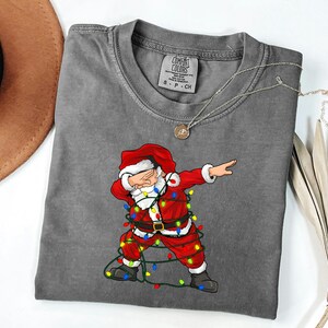May include: A charcoal gray t-shirt featuring a graphic of Santa Claus performing a dab dance move while wrapped in colorful Christmas string lights. The shirt has a crew neckline and a Comfort Colors brand label printed on the inside collar.