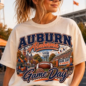 May include: A cream-coloured short-sleeve t-shirt featuring a graphic print with the text AUBURN ALABAMA GAME DAY. The design includes a football, a water tower, a stadium, and tailgating scenes in shades of blue, orange, and brown.