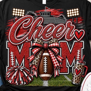 Op de afbeelding: Een zwart t-shirt met een grafische print met de tekst Cheer MOM in rode glitter. Het ontwerp bevat een football, een rood-zwarte pompon, een strik met luipaardprint en ruitjes, een stadionachtergrond en een beker met luipaardprint.