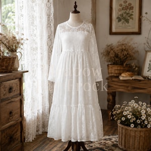 Op de afbeelding: Een witte jurk met lange mouwen, voorzien van een bloemmotief kanten overlay, een ronde halslijn en een rok met verschillende lagen. Het kledingstuk wordt getoond op een paspop en bevat de tekst Shadow Frock in het midden.