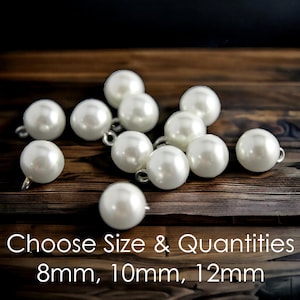 May include: A selection of glossy, white faux pearl beads with small loops, arranged on a dark wooden background. The image includes text that reads "Choose Size & Quantities" with size options of 8mm, 10mm, and 12mm.