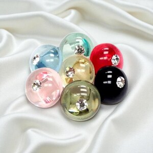 May include: A collection of seven colorful, round glass beads. Each bead is a different color, including red, black, pink, green, blue, and gold. A small, sparkling crystal is embedded in the center of each bead. The beads are arranged on a white, silky surface.