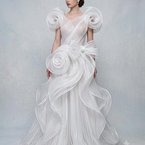 Puede incluir: Un vestido blanco largo hasta el suelo con escote en V, tela plisada transparente y grandes acentos escultóricos en espiral en los hombros y la cadera. El vestido incluye un lazo prominente en la cintura y capas de tela plisada voluminosas.