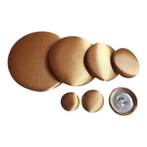 May include: A set of six gold-colored fabric covered buttons of varying sizes. The buttons are round and have a smooth, satin-like finish.