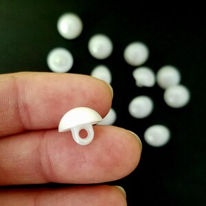 Puede incluir: Botones blancos en forma de hongo con un lazo en la parte inferior, dispersos sobre un fondo negro. Los botones son de material brillante. Un botón se sostiene entre los dedos.