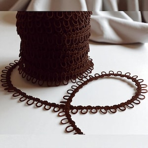 May include: A roll of dark brown looped trim with a decorative edge. The trim is unwound in a circular pattern, showing the detailed design. The background is a soft, neutral colour.