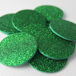 May include: A collection of round, green glitter embellishments. The circular shapes are scattered across a white surface, with the glitter catching the light. These decorative pieces could be used for crafts or other creative projects.