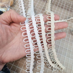 以下が含まれることがあります： 白とオフホワイトのループトリムのストリップのクローズアップ。トリムは手で持たれており、繊細で装飾的なデザインを示しています。トリムは、縫製、工芸、またはさまざまなプロジェクトへの装飾の追加に使用される可能性があります。