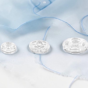 May include: Three clear plastic buttons of different sizes are displayed on a pale blue fabric with blue stitching. The buttons have a circular design with small holes around the centre.