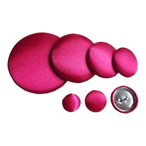 May include: A collection of magenta fabric-covered buttons in various sizes. The buttons range from large, flat circles to smaller, rounded shapes, with one button showing a metal back with buttonholes. The fabric has a subtle texture.