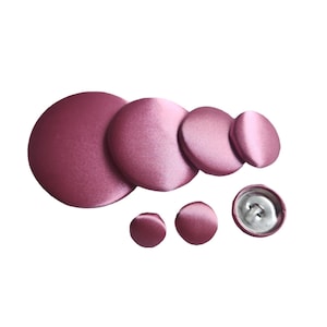 May include: Assortment of six pink satin buttons in various sizes. The buttons are round and have a smooth, shiny finish.