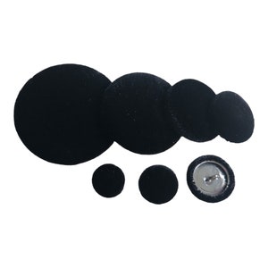 May include: A collection of black velvet covered buttons in various sizes. The buttons range from small to large, with one button showing a metal shank on the back. The buttons are arranged on a white background.