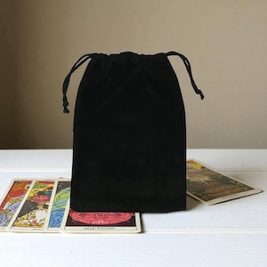 Può includere: Un sacchetto nero in velluto con coulisse, probabilmente per carte dei tarocchi, è appoggiato su una superficie bianca. Sono visibili diverse carte dei tarocchi, con illustrazioni colorate e testo. Il sacchetto è chiuso con un cordoncino nero.