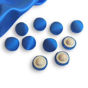 May include: A collection of eight cobalt blue fabric-covered buttons, some showing their metal and fiber backing. A piece of matching blue fabric is in the upper left corner. The buttons are round and have a smooth, satin-like texture.
