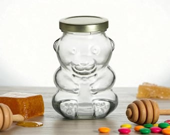 May include: A clear glass bear-shaped honey jar with a gold-colored lid. The jar is sitting on a wooden surface with a wooden honey dipper, a block of honey, and colorful candies.