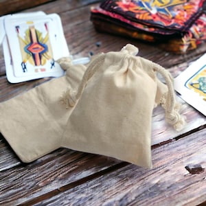 May include: Two small, off-white, drawstring fabric bags made of cotton. The bags are lying on a wooden surface.