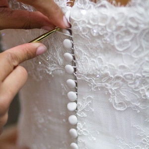May include: Close-up of a white lace wedding dress being buttoned. The dress features delicate lace detailing and small, round buttons. A gold crochet hook is used to fasten the buttons, highlighting the intricate design and craftsmanship of the bridal gown.