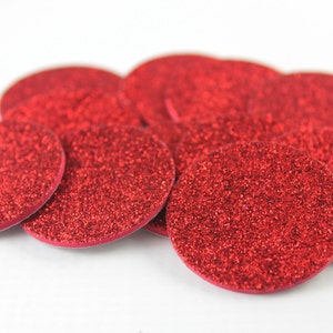 May include: A collection of red glittery, circular embellishments. The embellishments are scattered across a white surface. The glitter adds a sparkling effect to the red circles. These could be used for crafts or decorations.