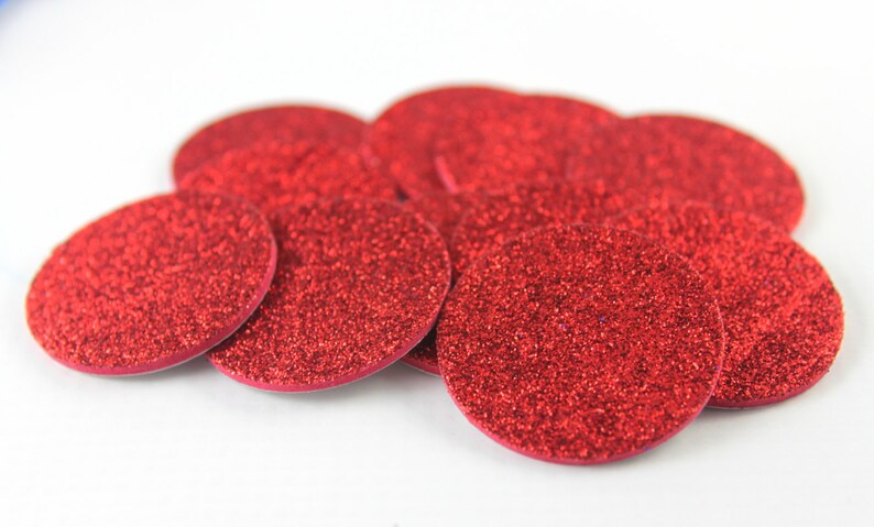 May include: A collection of red glittery, circular embellishments. The embellishments are scattered across a white surface. The glitter adds a sparkling effect to the red circles. These could be used for crafts or decorations.