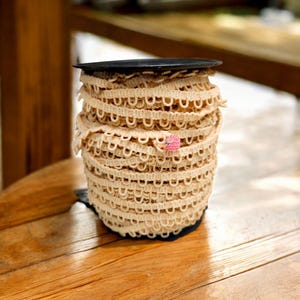 May include: A spool of beige elastic trim with a decorative scalloped edge. The trim is wound around a black plastic spool.