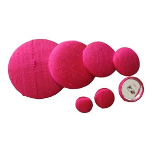 May include: A set of seven pink fabric covered buttons of various sizes. The buttons are round and have a smooth, slightly textured surface.