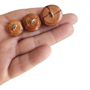 May include: Three brown leather buttons with metal loops. The buttons are different sizes and are arranged in a row from largest to smallest.