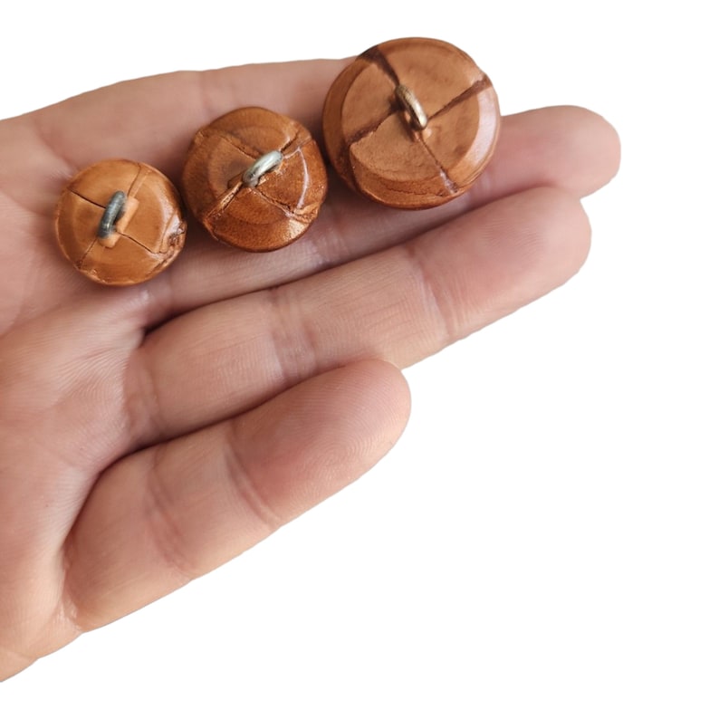 May include: Three brown leather buttons with metal loops. The buttons are different sizes and are arranged in a row from largest to smallest.