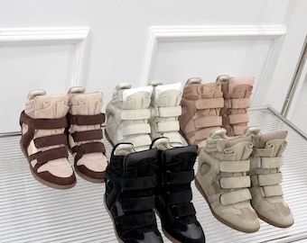 Könnte beinhalten: Eine Sammlung von vier Paar hohen Keil-Sneakern, die auf einem Glasregal angeordnet sind. Die Sneaker verfügen über mehrere Klettverschlüsse auf der Vorderseite, gepolsterte Kragen sowie eine Konstruktion aus Wildleder und Leder in Schwarz, Beige, Hellbraun und Braun.