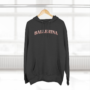 May include: A charcoal gray hooded sweatshirt featuring a front kangaroo pocket, long sleeves, and a drawstring hood. The word BALLERINA is printed in a curved, metallic rose gold font across the center of the chest on this long-sleeved pullover.