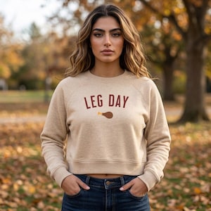Puede incluir: Una sudadera beige de manga larga y cuello redondo con el texto LEG DAY en letras de color rojo oscuro en el pecho, acompañado de un pequeño gráfico de un muslo de pavo asado de color marrón situado debajo del texto.