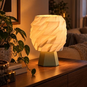 Pode incluir: Um candeeiro de mesa com um abajur branco geométrico facetado que emite uma luz quente e suave. O candeeiro tem uma base hexagonal sólida em cor verde sálvia, proporcionando uma base estável para a estrutura angular e texturizada.