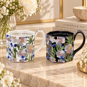 Può includere: Due tazze in ceramica ottagonali, una bianca e una nera, con un colorato motivo floreale di foglie blu, rosa e verdi. Ogni tazza è dotata di manico e della scritta "NATURAL SERENITY AND BEAUTY" stampata lungo il bordo interno superiore.